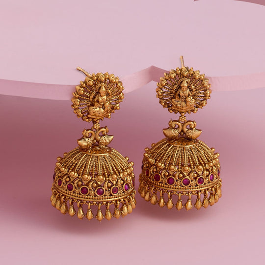 Antique jhumka earrings with peacock design, ruby-red stones, on pink background.