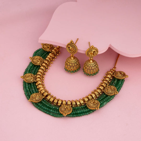 Antique necklace and earrings with green beads and peacock motifs on pink surface.