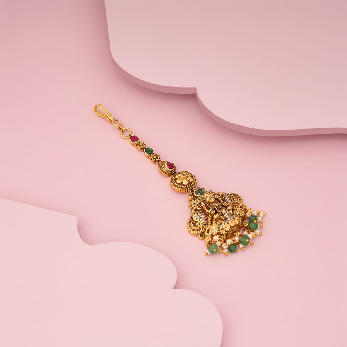 Antique tikka with deity motif, green stones, on pink surface.