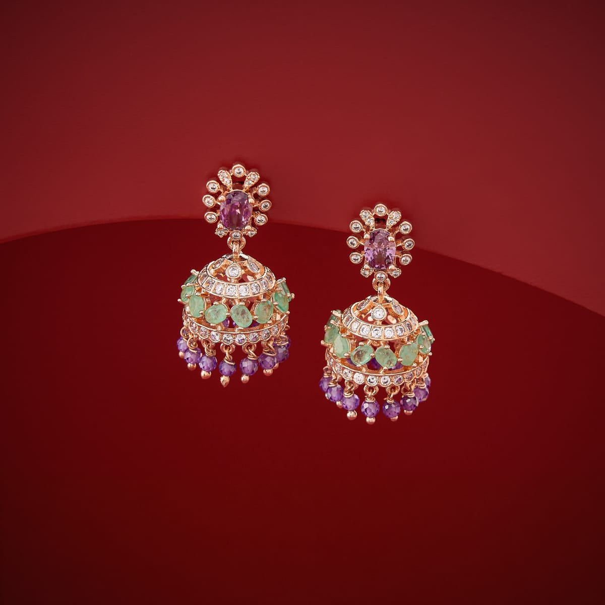 Kundan jhumka earrings with green and lavender stones, gold polish, on red surface.