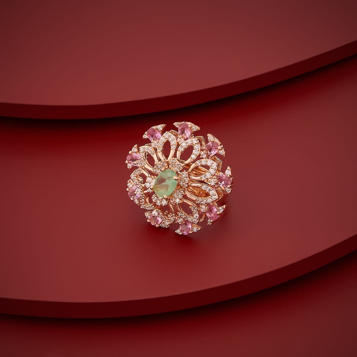 Gold-tone finger ring with green center stone and pink accents, angled view.