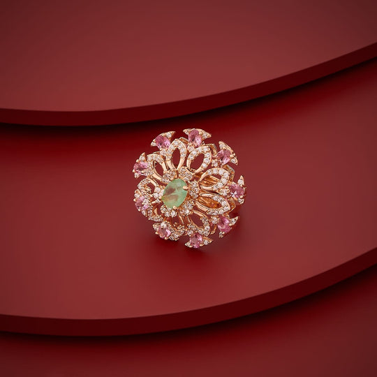 Gold-tone finger ring with green center stone and pink accents, angled view.