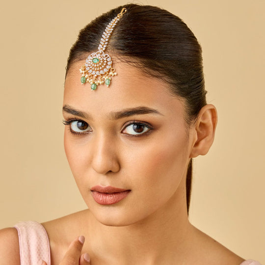 Model wearing floral zircon tikka with green and white stones, gold polish.