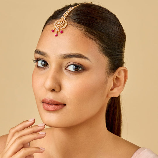 Model wearing gold polish zircon tikka with ruby drops, floral motif.